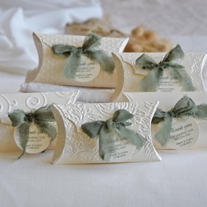 Puede incluir: Cajas en forma de almohada de color crema con diseños en relieve y lazos de cinta verde salvia. Cada caja tiene una etiqueta que dice "Thank you for sharing our special day". Ideales para recuerdos de boda.