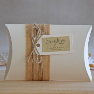 May include: White pillow box with a brown burlap ribbon and a tag that says "from us to you" with the names Martha & Hugh and the date November 24th, 2021.