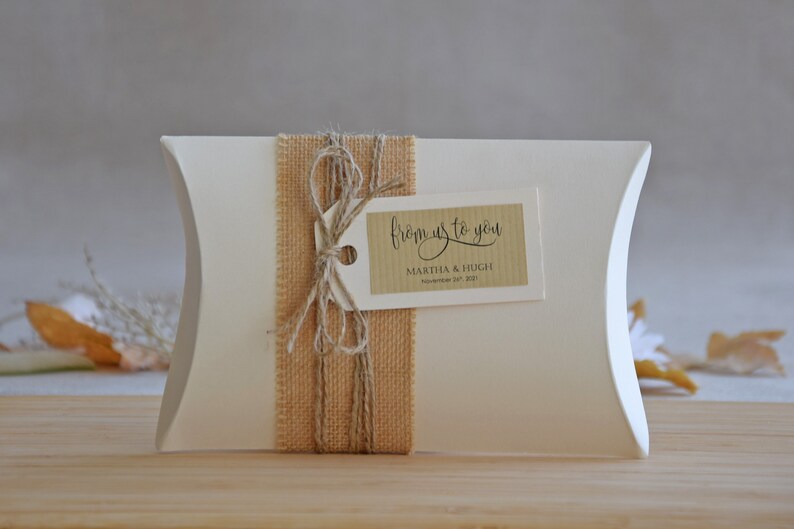 May include: White pillow box with a brown burlap ribbon and a tag that says "from us to you" with the names Martha & Hugh and the date November 24th, 2021.