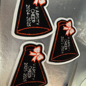 May include: Three embroidered patches in the shape of a megaphone. Each patch is black with orange trim and a white bow at the top. The text "VARSITY CHEER" and a year range are embroidered in white.