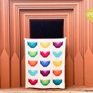May include: A colorful quilt with a repeating floral pattern in shades of blue, green, red, purple, and yellow. The quilt is displayed against a rustic, orange-toned wooden backdrop. A PDF icon is visible in the upper right corner.