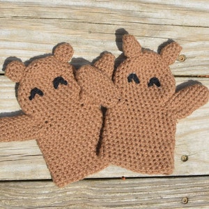 May include: Two brown crocheted bear hand puppets with black eyes. The puppets are on a wooden surface.