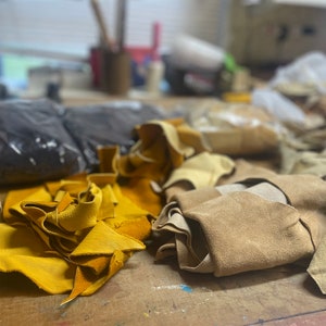 May include: A collection of leather scraps in various colors, including yellow, tan, and black, are scattered across a wooden surface. The leather pieces appear to be of different sizes and shapes, suggesting they are remnants from a crafting project. The background is blurred.