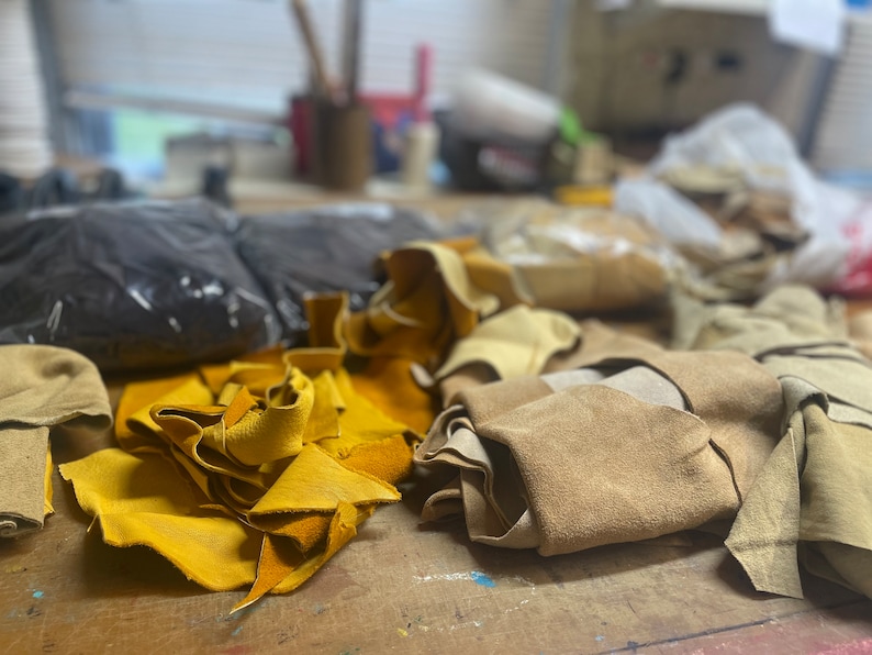 May include: A collection of leather scraps in various colors, including yellow, tan, and black, are scattered across a wooden surface. The leather pieces appear to be of different sizes and shapes, suggesting they are remnants from a crafting project. The background is blurred.