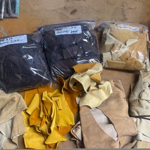 May include: Assortment of leather scraps in various shades of brown and tan. Three bags labeled "Brown Deer", "Chocolate Deer", and "Smoke Deer" are visible. The leather pieces are of different sizes and textures, suitable for crafting.