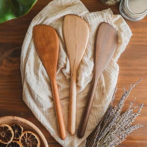 May include: Three wooden cooking spatulas with smooth, rounded handles and wide, flat heads. Each spatula features a distinct wood grain and color, ranging from light beige to medium brown and a deeper, darker brown tone.