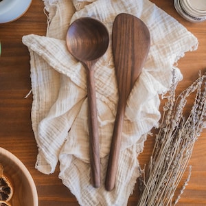 May include: Wooden kitchen utensils, a ladle and a spatula, rest on a cream-colored cloth. The utensils are a rich brown color and appear to be made of hardwood. Dried lavender and a wooden bowl with dried citrus slices are also visible.