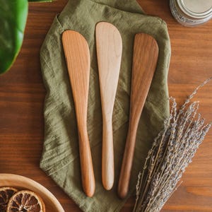 May include: Three wooden spreaders with smooth, rounded handles and flat, curved blades. Each spreader features a distinct wood grain pattern and color, ranging from light beige to medium brown, arranged vertically on a textured olive green cloth.
