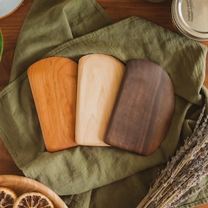 May include: Three wooden dough scrapers arranged in a row, featuring distinct wood tones ranging from a light cream to a medium reddish-brown and a dark espresso brown. Each tool has a smooth, curved edge and a flat, rectangular base.