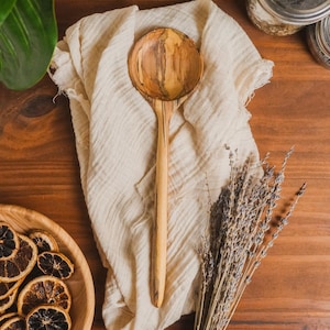 May include: A handcrafted wooden spoon featuring a round bowl and a long, slender handle with natural wood grain patterns in shades of light tan and dark brown, displayed on a textured off-white fabric cloth.