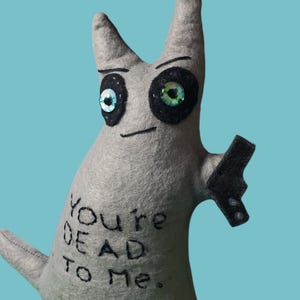 May include: A gray felt creature with large, expressive eyes and a small, black gun. The text "You're DEAD TO Me." is embroidered on the body. The background is a solid teal color.