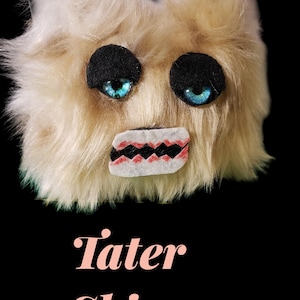 May include: A plush, furry creature with light tan fur, black eyes, and blue irises. It has a stitched mouth with red and white detailing. The text "Tater Skinz" is displayed below the creature.