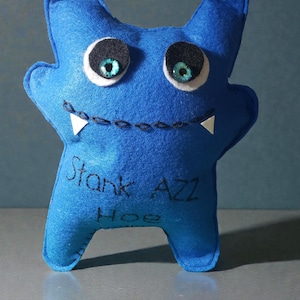 May include: A blue felt monster plushie with large eyes, pointed ears, and a stitched smile. The plushie has the words "Stank AZZ Hoe" written on it.