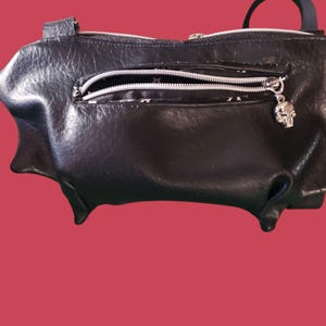 May include: Black bat-shaped handbag with a silver zipper and a skull charm. The bag has a textured appearance and a small handle. The background is a solid red color.