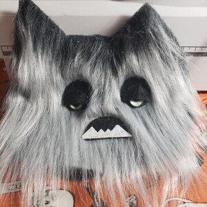 May include: A plush, furry monster face with black and gray fur, black eyes, and a white, jagged mouth. The monster has pointed ears and a grumpy expression. The background is an orange fabric with Halloween-themed patterns.