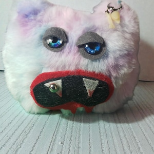 May include: A plush, abstract creature with a fluffy, multi-colored body in shades of white, pink, and blue. It has large, blue eyes with gray eyelids, a red mouth, and a black snout with silver and green accents. A small, dangling charm is attached to its ear.