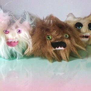 May include: Three handmade plush monster toys with shaggy fur in pastel colors. Each has large, gem-like eyes and a stitched mouth. The monsters are arranged on a gradient background of pink and green.