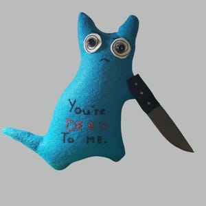 May include: A handmade blue felt creature with large, expressive eyes and a sad expression. It holds a black and silver knife. The text "You're DEAD To ME." is embroidered in red on its body. A unique, quirky plush toy.