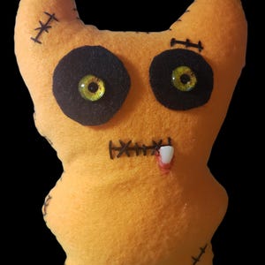 May include: Orange plush monster with black stitched details, including eyes, mouth, and stitches. The monster has yellow and black eyes and a single white tooth. The toy is handmade and has a stitched design.