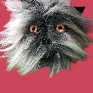 May include: A plush creature head with a furry texture in shades of gray and white, featuring large, detailed eyes with yellow and orange hues. The creature has black pointed ears and a small, triangular tag.
