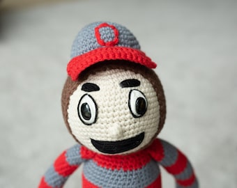 May include: A crocheted plush toy wearing a red and gray baseball cap with a white "O" on it. The toy has a red and gray striped shirt and a white face with black eyes and a smile.