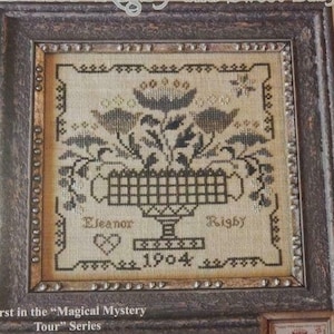 May include: A cross-stitch pattern featuring a floral arrangement in a basket with the text "Eleanor Rigby and Sweet Baby" and "1904". The pattern is framed in a dark wood frame with a distressed finish. The pattern is part of the "Magical Mystery Tour" series.