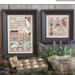 May include: A framed cross-stitch sampler with the text "April brings the Primrose Sweet & scatters Daisies at our Feet" and a matching smaller sampler. A decorative bunny statue and a box of eggs are also present. The word "APRIL" is at the bottom.