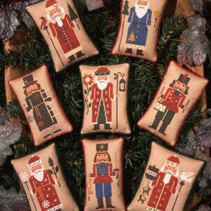 May include: Eight embroidered pillow ornaments featuring different nutcracker designs. Each pillow is beige with a brown trim and features a different nutcracker character in a colorful embroidered design.