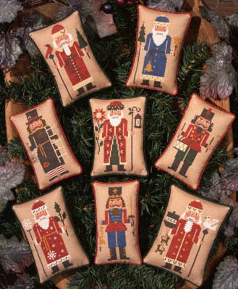May include: Eight embroidered pillow ornaments featuring different nutcracker designs. Each pillow is beige with a brown trim and features a different nutcracker character in a colorful embroidered design.