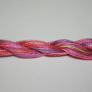 May include: A close-up of a braided strand of embroidery floss. The floss is a blend of pink, red, purple, and yellow hues. The thread is tightly twisted, creating a textured appearance. The background is a plain, light gray.