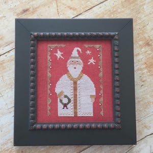 May include: Framed cross-stitch art featuring Santa Claus in a white robe with gold trim, holding a wreath. The artwork is set against a red background with white stars and a beaded border, framed in a dark gray frame.