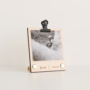 May include: A wooden photo frame with a black clip holding a black and white photo. The frame has a brass stand and the text "Jacob | 25.01.23" is engraved on the bottom.