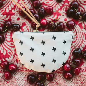 May include: A small, white canvas pouch with a black Texas state outline pattern. The pouch has a gold zipper and a leather pull. The pouch is surrounded by fresh cherries on a red and white patterned surface.