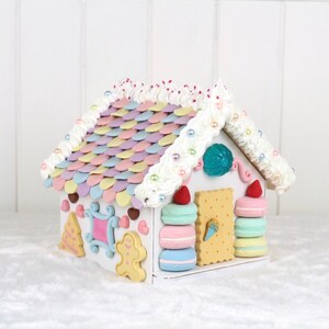 May include: A white gingerbread house decorated with pastel-colored candy, frosting, and pearl beads. The roof is made of pastel-colored candy shingles. The house has a gingerbread man, a Christmas tree, and a window with a heart design.