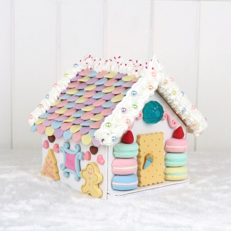 May include: A white gingerbread house decorated with pastel-colored candy, frosting, and pearl beads. The roof is made of pastel-colored candy shingles. The house has a gingerbread man, a Christmas tree, and a window with a heart design.