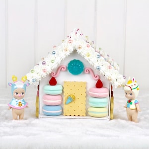 May include: A white gingerbread house decorated with pastel-colored macarons, candy, and pearl beads. Two small figurines stand on either side of the house.