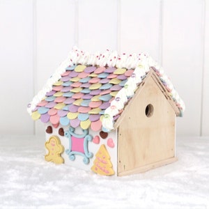 May include: A pastel-colored gingerbread house with a white frosting roof and candy decorations. The house has a gingerbread person, a heart, and a Christmas tree on the front.