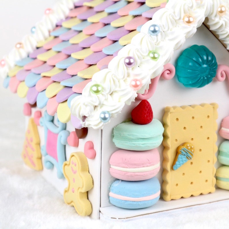 May include: A miniature gingerbread house decorated with pastel-colored frosting, macarons, and a strawberry. The roof is made of colorful candy shingles and the house is trimmed with white frosting and pearl sprinkles.