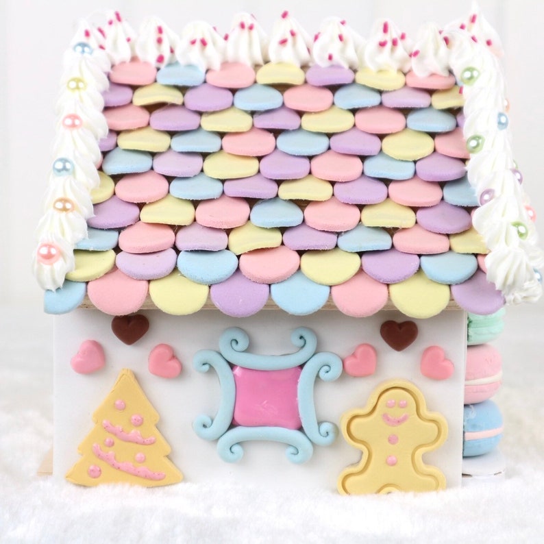May include: A gingerbread house decorated with pastel-colored frosting and candy. The roof is covered in pastel-colored candy hearts. The house has a gingerbread tree, a gingerbread man, and a gingerbread window.
