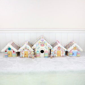 May include: Five miniature gingerbread houses decorated with pastel-colored candies and frosting. Each house has a unique design and features a small doll figurine standing in front of it.