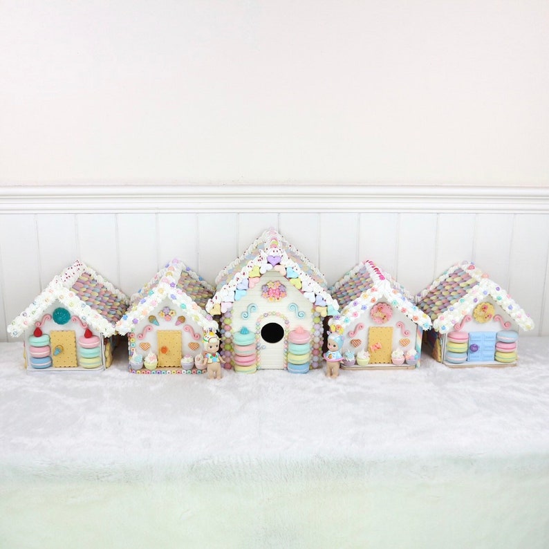 May include: Five miniature gingerbread houses decorated with pastel-colored candies and frosting. Each house has a unique design and features a small doll figurine standing in front of it.