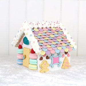 May include: A white gingerbread house decorated with pastel-colored candy, white frosting, and pearl-like sprinkles. The roof is made of candy shingles and the house has a gingerbread door and windows.