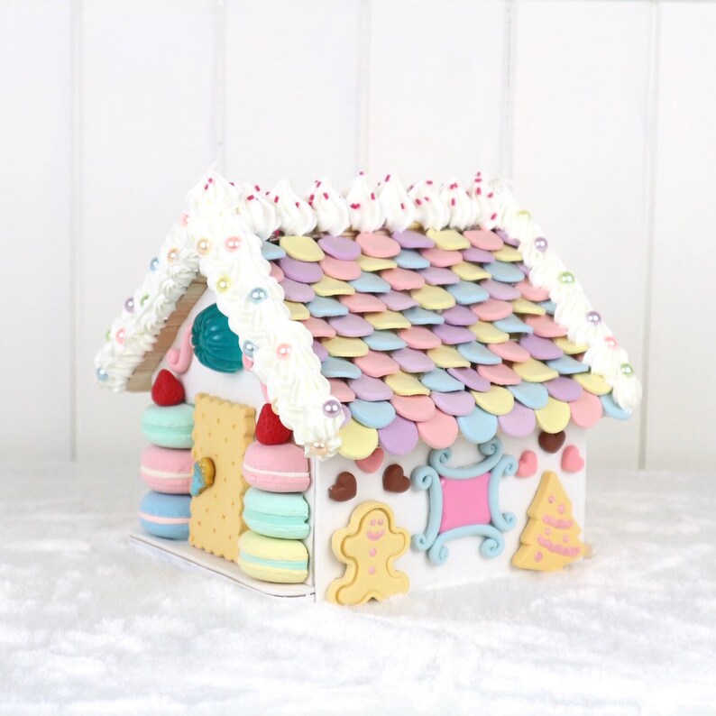 May include: A white gingerbread house decorated with pastel-colored candy, white frosting, and pearl-like sprinkles. The roof is made of candy shingles and the house has a gingerbread door and windows.