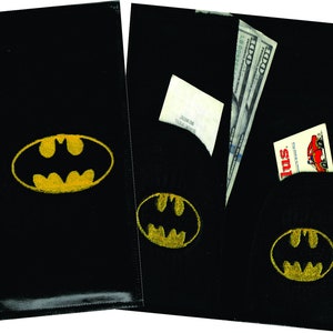May include: Two black leather wallets with a yellow Batman logo. The wallet on the right has a pocket with a dollar bill and a small card sticking out.