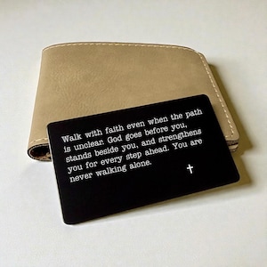 May include: A tan leather wallet with a black card in front. The card has white text that reads, "Walk with faith even when the path is unclear... You are never walking alone." A white cross is at the bottom.