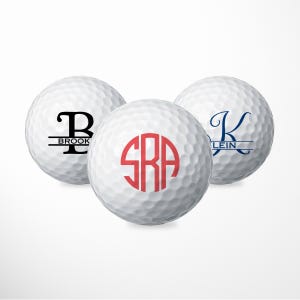 May include: Three white golf balls with personalized monograms. The first ball has a black monogram "B" with the name "BROOK" underneath. The second ball has a red monogram "SRA". The third ball has a blue monogram "K" with the name "KLEIN" underneath.