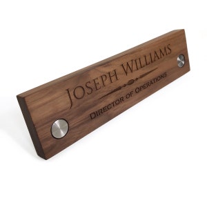 May include: A wooden desk nameplate with the text "Joseph Williams, Director of Operations" engraved in a dark font. The nameplate has two silver metal accents on the sides.