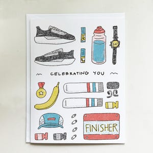 May include: A white card with a hand-drawn illustration of running shoes, a water bottle, a watch, and other athletic accessories. The text "CELEBRATING YOU" is written across the center of the card. The word "FINISHER" is written on a red sign.