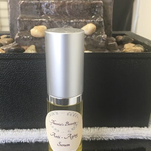 May include: A small, clear glass bottle with a silver cap, filled with a yellow liquid. The bottle has a label that reads "Theoria's Beauty Anti-Aging Serum". The background includes a small decorative fountain.