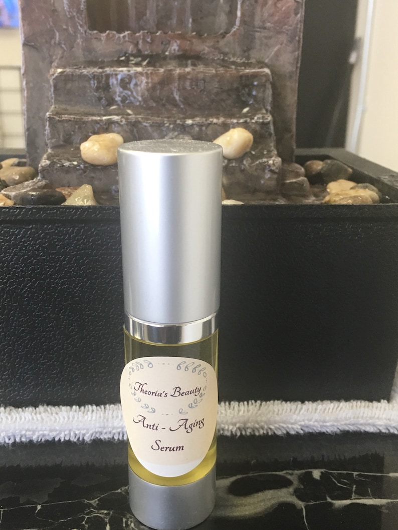 May include: A small, clear glass bottle with a silver cap, filled with a yellow liquid. The bottle has a label that reads "Theoria's Beauty Anti-Aging Serum". The background includes a small decorative fountain.